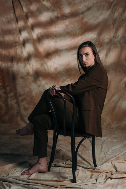 Full length of brunette queer person in suit sitting on chair on abstract brown background - Φωτογραφία, εικόνα