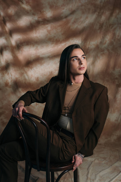 Trendy nonbinary person looking away while sitting on chair on abstract brown background - Fotó, kép