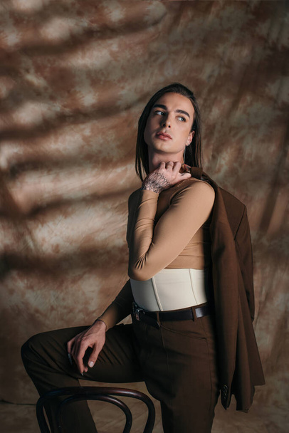 Nonbinary person in corset holding jacket near chair on abstract brown background - Foto, imagen