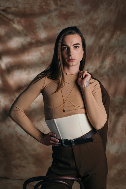 Fashionable queer person in corset holding blazer on abstract brown background - Zdjęcie, obraz