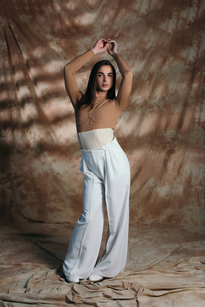 Stylish nonbinary person in corset and white pants standing on abstract brown background  - Fotografie, Obrázek
