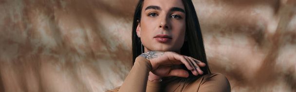 Portrait of young tattooed queer person looking at camera on abstract brown background, banner  - Photo, image