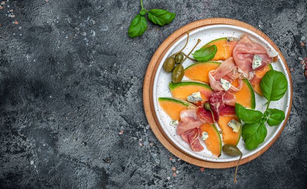 Salad with fresh melon with prosciutto and basil on a dark background. Antipasti, Traditional Spanish and Italian appetizer. Long banner format. top view. - Photo, Image
