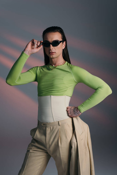 Brunette nonbinary model in corset and sunglasses holding jacket on abstract background  - Foto, afbeelding