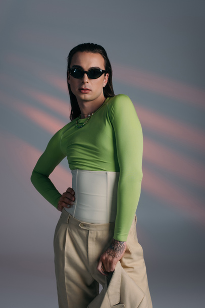 Fashionable nonbinary model in sunglasses holding jacket on abstract background  - Fotografie, Obrázek