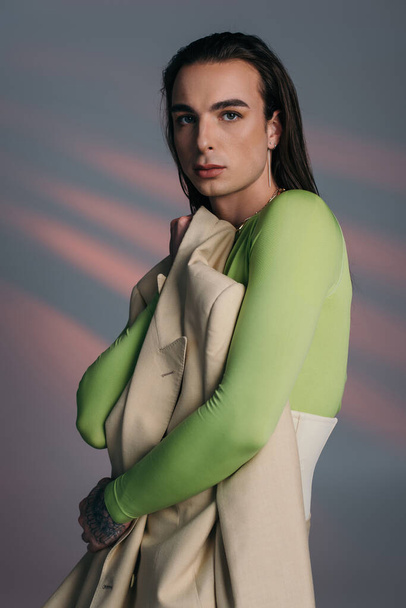 Brunette queer model holding jacket and looking at camera on abstract background  - Zdjęcie, obraz