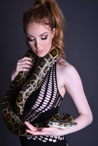 Beautiful, tall, redhead model in a black dress, with her pet python, against a dark grey background - Foto, imagen