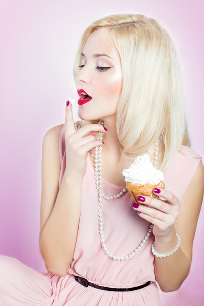 Beautiful sexy elegant blonde woman with bright makeup in pink dress in the Studio on a white background sitting with a cupcake in her hands and licks the sweet cream from his finger winks retro style - Foto, Imagem