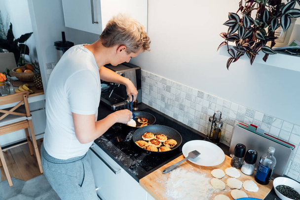 A young man is cooking cheese pancakes in a modern light kitchen at home. Man preparing pancakes for frying on the pan. Housework and home duties. Culinary hobby.High view. Selective focus - Foto, Imagen