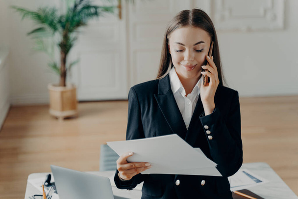 Smiling attractive young businesswoman in formal suit talking on phone and looking at notes, holding documents and discussing business ideas on smartphone, female manager standing in office - Foto, Imagen