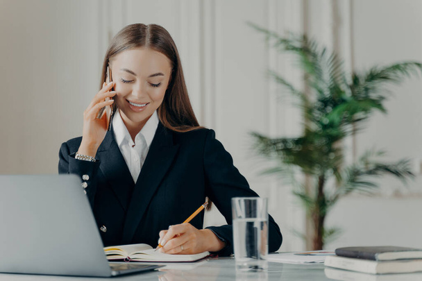 Young experienced business consultant in elegant black suit talking on phone, writing notes with pencil in notepad while communicating with client, female manager working on laptop at her workplace - Photo, Image
