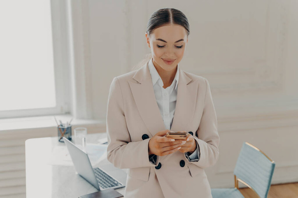 Glücklich schöne Unternehmensberaterin in formaler Kleidung mit Handy, Nachrichten lesen oder SMS senden, während sie drinnen in einem modernen hellen Büro steht und geschäftigen Arbeitstag genießt - Foto, Bild