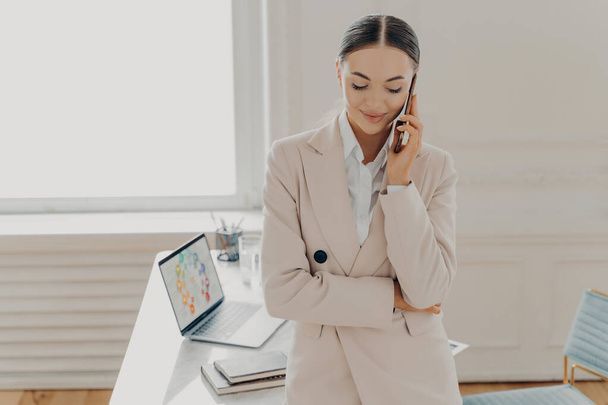 Busy female director in formal suit having cell phone conversation, talking on phone and discussing new project with copartner. Businesswoman working at light office with laptop in wooden desk - Фото, зображення