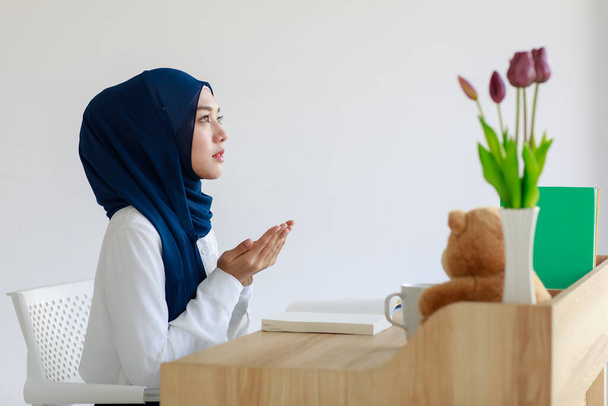 Красивая молодая азиатская мусульманка в красивом хиджабе и платке поднимает руки в молитве. Женщина исламская студентка молится во время чтения книги. Концепция веры и обучения. - Фото, изображение