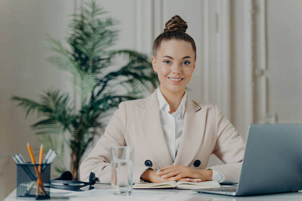 Portret van een mooie, zelfverzekerde zakenvrouw in elegante kleding die aangenaam glimlacht terwijl ze op kantoor zit en moderne technologieën gebruikt, werkend op een laptop. Functie- en beroepsconcept - Foto, afbeelding