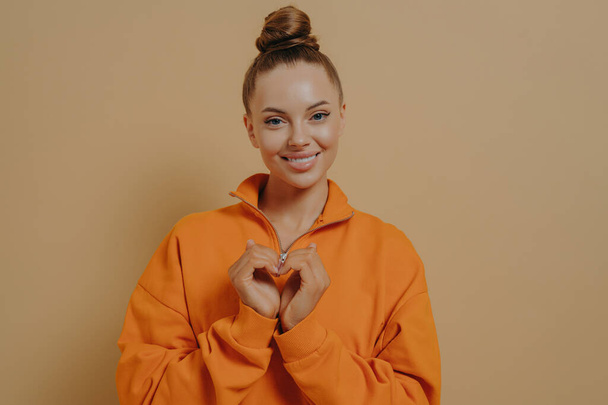 Share love. Smiling grateful young woman making heart gesture, expressing warm feelings to boyfriend, isolated on beige studio background, female volunteer showing heart sign, Charity concept - Foto, afbeelding