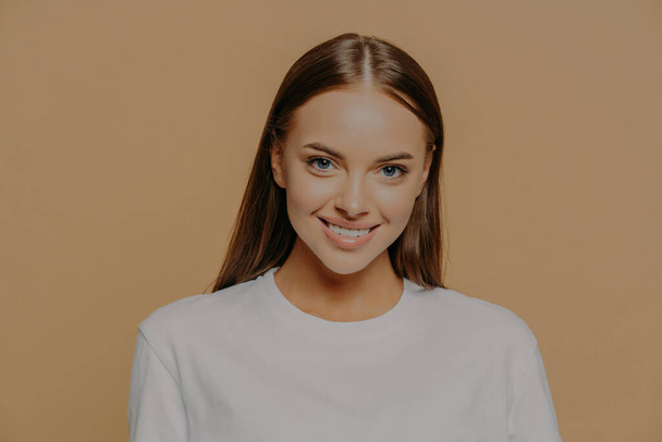 Portrait of beautiful woman with long dark straight hair smiles gently wears casual white jumper has minimal makeup poses against brown background. Women beauty and positive emotions concept - Photo, Image