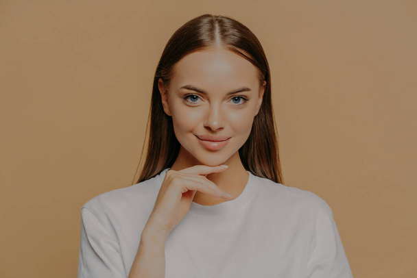Lovely young European woman with makeup healthy skin touches gently jawline looks directly at camera has long straigh hair natural beauty dressed in casual sweater isolated over brown background - Foto, Bild