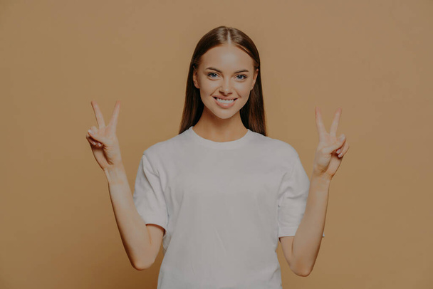 Horizontal shot of happy optimistic young Caucasian woman raises arms shows peace gesture with fingers does victory sign says number two wears casual white t shirt isolated over brown background - Photo, Image