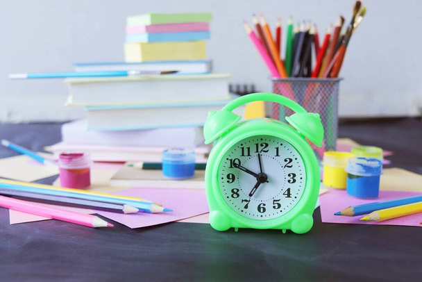Будильник на столе, на фоне разноцветной бумаги, книг и канцелярских принадлежностей, обратно в школу, образование, стол для школьников - Фото, изображение