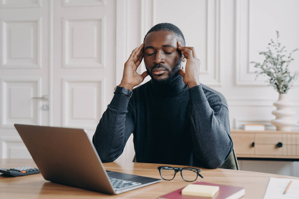 Frusztrált fáradt afro-amerikai férfi irodai dolgozó csukott szemmel próbál koncentrálni, miközben ül a munkahelyen, afrikai etnikai férfi alkalmazott masszírozó templomok, szenved fejfájás a munkahelyen - Fotó, kép