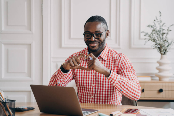 Молодой счастливый романтичный африканский мужчина смотрит на ноутбук во время видеозвонка с подругой, показывает символ сердца с руками и пальцами, веселый работник разговаривает с любимым человеком во время удаленной работы в Интернете - Фото, изображение