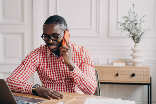 Młody, nowoczesny afrykański biznesmen rozmawiający przez telefon komórkowy z klientem lub partnerem podczas pracy zdalnej w domu, zadowolony mężczyzna afro amerykański pracownik biurowy wykonujący połączenie za pomocą smartfona w pracy - Zdjęcie, obraz