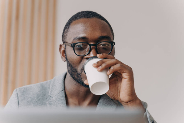 Attractive confident Afro american man company CEO drinking take away coffee from disposable cup while looking at laptop screen following remote financial meeting online during video conference - Foto, Imagem