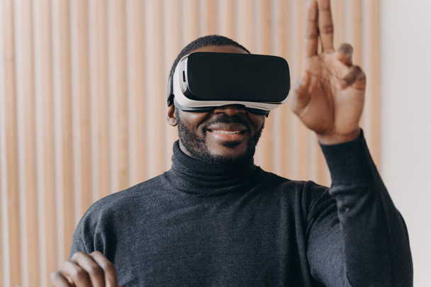 Aufgeregte Afroamerikaner mit VR-Headset-Brille genießen Augmented Reality, während sie zu Hause am modernen Arbeitsplatz sitzen, fröhlich erhobene Hand mit zwei erhobenen Fingern als Interaktion mit dem Cyberspace in 3D - Foto, Bild