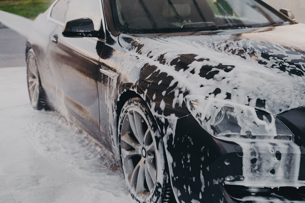 Washing off white soap foam with water from luxury black car at self-service car wash station outdoors, using high pressure washer and detergent for professional vehicle cleaning - Fotoğraf, Görsel