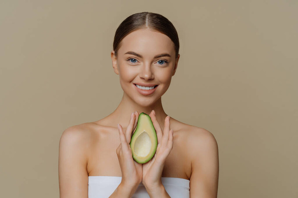 Beauty and personal care concept. Smiling calm European woman with combed hair holds avocado as recommendation for nourishing face mask wrapped in bath towel isolated over beige studio background - Foto, Bild