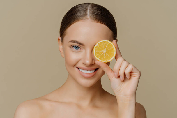 Beauty and vitamin C concept. Headshot of lovely dark haired European woman smiles gently covers eye with slice of juicy lemon poses half naked against brown wall has healthy skin eats healthy food. - Foto, afbeelding
