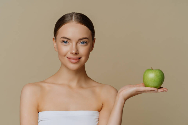 Photo of healthy lovely woman with natural makeup stand wrapped in bath towel holds green apple recommends product for dieting isolated over brown background makes natural homemade facial masks - Φωτογραφία, εικόνα