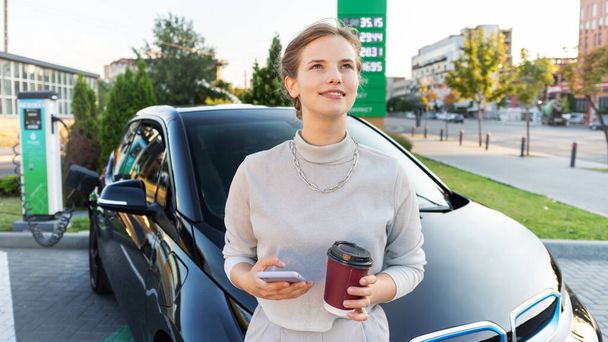 A young blonde woman with smartphone and coffee at a car charging station with charging electric car nearby in Chisinau, Moldova - Foto, afbeelding