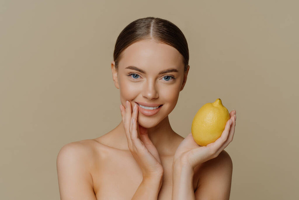 Portrait of charming half naked woman with dark combed hair holds fresh whole lemon uses fruit for natural cosmetics poses against brown background with naked body. People beauty wellness concept - Foto, Bild