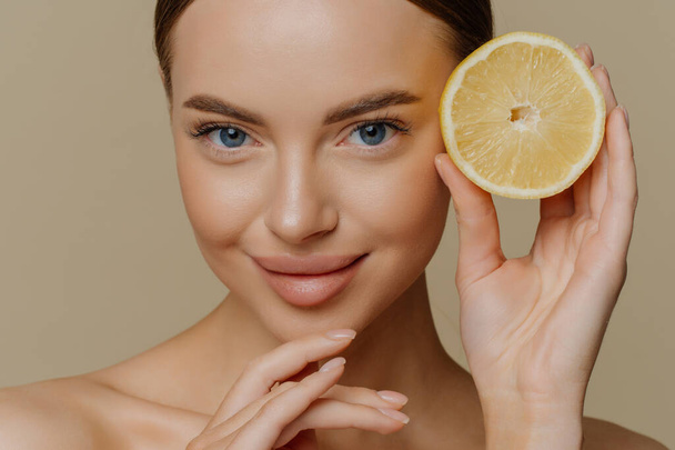 Beauty portrait of healthy young topless brunette woman touches chin gently holds half of juicy fresh lemon has natural makeup poses topless isolated over brown background. Facial care concept - Photo, Image