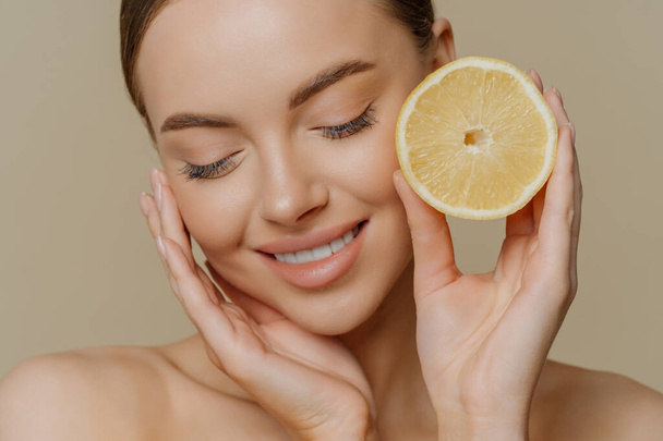Headshot of sensual charming young woman wears minimal makeup touches face gently holds slice of lemon cares about freshness of skin has citrus peeling stands half naked against brown background - Foto, Bild