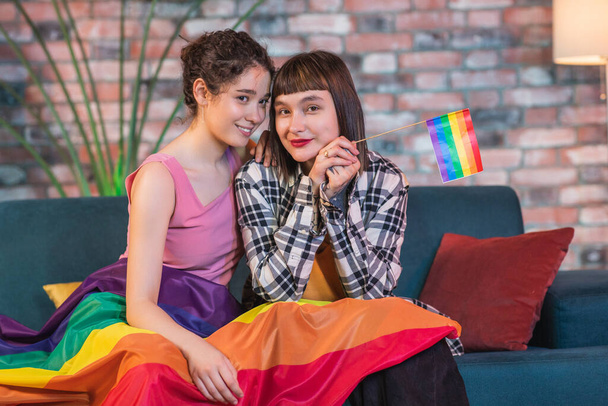 In living room on the sofa closeup to the camera very lovely lesbian couple hugging closeup each other and holding in hands LGBT rainbow flag. - Foto, Imagem