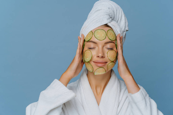 Beauty skin care anti aging concept. Relaxed European woman undergoes beauty treatments uses natural cosmetology keeps eyes closed wears dressing gown and wrapped towel on head models indoor - Zdjęcie, obraz