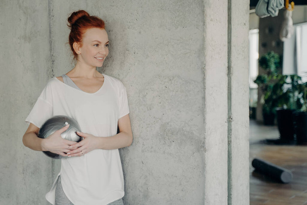 Zadowolona rudowłosa kobieta w koszulce whire stojąca pod betonową ścianą z małym fitballem w ręku, ciesząca się czasem treningowym w studiu fitness, spoglądająca na bok i uśmiechnięta. Koncepcja zdrowego stylu życia - Zdjęcie, obraz