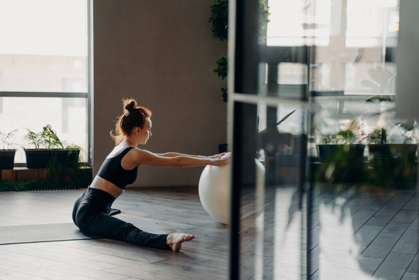 Widok przez otwarte szklane drzwi elastycznej rudej szczupłej kobiety w aktywnym zużyciu siedzi w sznurku na macie z dłońmi leżącymi na dużym fitball podczas wykonywania ćwiczeń rozciągających w siłowni lub studio fitness - Zdjęcie, obraz