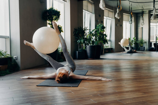 Sportieve vrouw liggend op yogamat met uitgestrekte armen die een oefenbal tussen de benen vasthoudt terwijl ze traint in een moderne fitnessstudio met grote spiegel en stabiliteitsoefeningen doet. Pilates concept - Foto, afbeelding