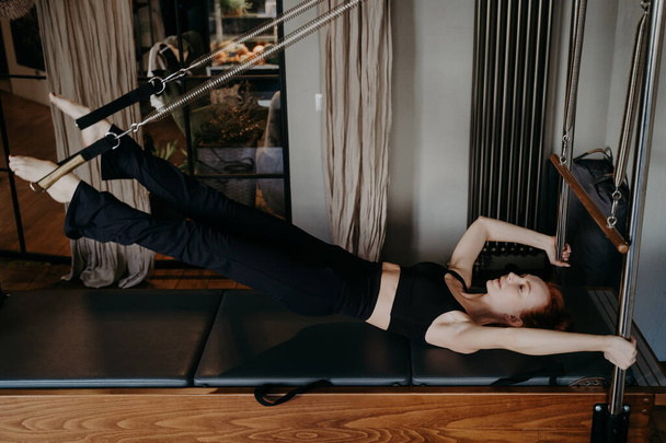 Fitness stüdyosunda sıkı çalışma. Motive olmuş genç fit kadın pilates eğitmeni Cadillac reformcusu üzerinde güç egzersizi yapıyor, her kas hareketinde güç gösteriyor. Aktif ve sağlıklı yaşam tarzı - Fotoğraf, Görsel