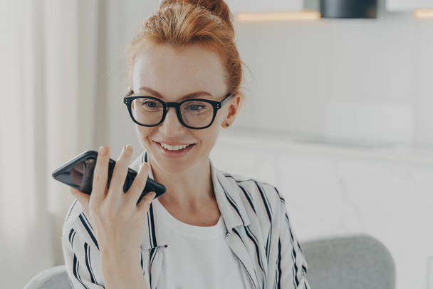 Close-up portrait of beautiful positive red-haired woman in spectacles holding mobile phone and sending audio message to friend while sitting in whote kitchen at home, using speakerphone loudspeaker - Foto, Bild