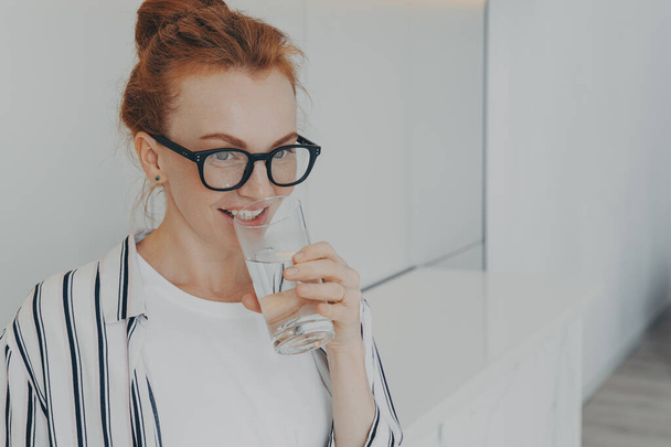 Happy healthy beautiful young redhead woman with clean skin drinking water in morning and smiling, beautiful female holding glass in hand, preventing dehydration while standing in kitchen at home - Foto, Imagem