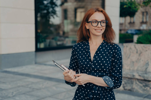 Ευτυχισμένη χαμογελαστή κοκκινομάλλα επιχειρηματίας κρατώντας φορητό υπολογιστή και σημειωματάριο, κοιτάζοντας στην άκρη, ενώ στέκεται στο δρόμο της πόλης σε εξωτερικούς χώρους. Γυναικεία επαγγελματική επιχείρηση με ψηφιακή συσκευή κοντά στο κτίριο γραφείων - Φωτογραφία, εικόνα