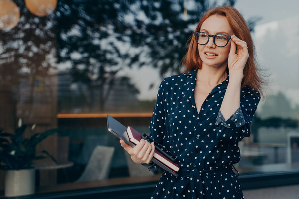 Venkovní záběr ženského podnikatele se dívá pozorně do dálky udržuje ruku na brýlích nosí černé puntíkové šaty nese tablet smartphone a organizátor pózuje v blízkosti okna restaurace - Fotografie, Obrázek