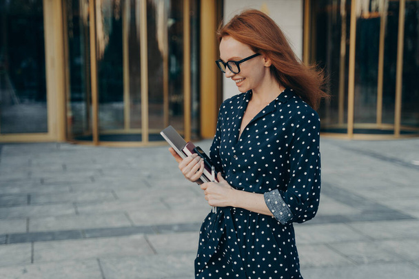 Happy ginger successful female executive worker strolls outdoor looks down happily wears spectacles polka dot dress holds laptop notebook and smartphone thinks about something has successful day - Foto, Imagem