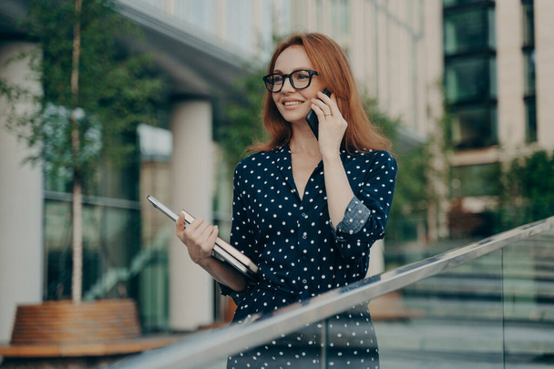 Happy adult lady with red hair makes consultancy call on smartphone spends leisure for international talking wears spectacles polka dot dress holds diary poses outdoor in urban setting focused forward - Photo, image