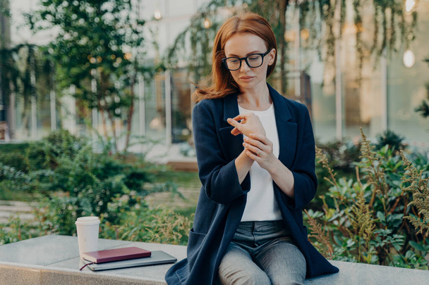 Thoughtful displeased redhead young woman suffers from pain in wrist touches hand sits outside on bench with disposable cup of coffee notebook digital device wears formal clothes focused down - Foto, afbeelding